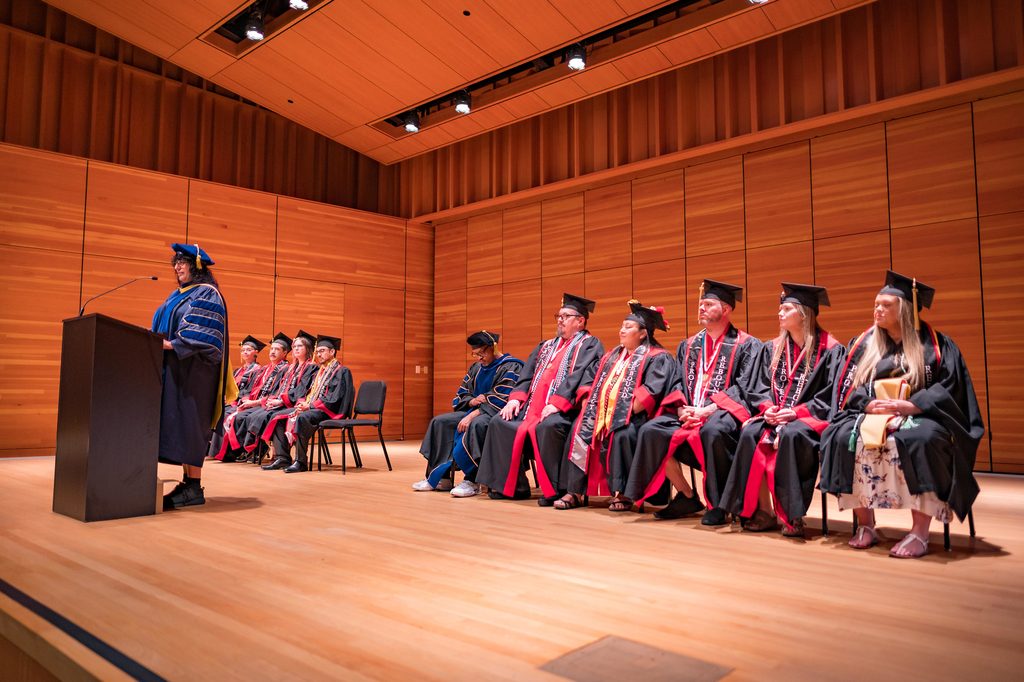 Woman speaking in front of an audience with recent graduates behind her.